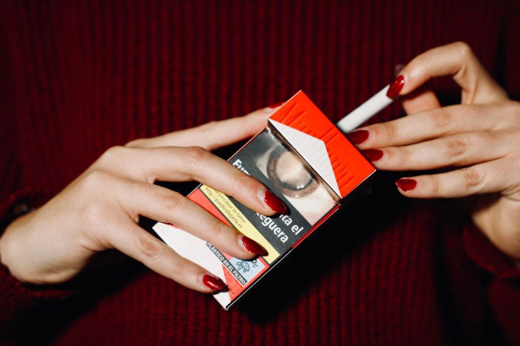 Close-up of hands with red nails holding a cigarette pack indoors.