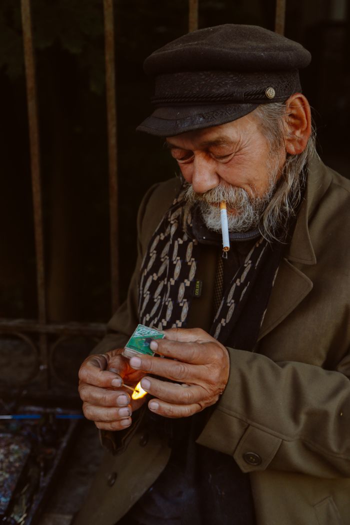 Senior ethnic bearded male in casual clothes and hat lighting cigarette with match while sitting near aged rusty fence on city street