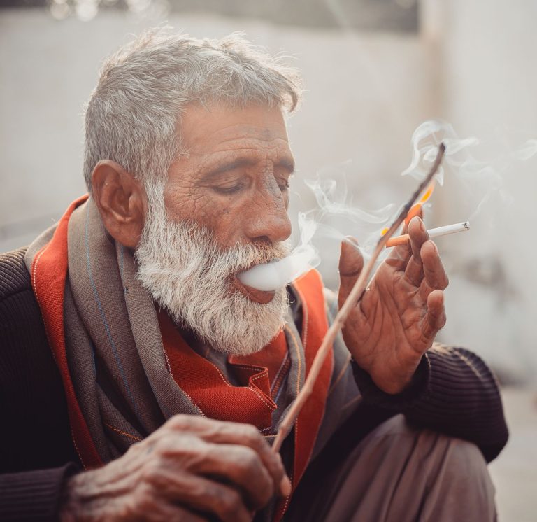 Old bearded ethnic male with wooden stick exhaling puffs of smoke while smoking cigarette on street