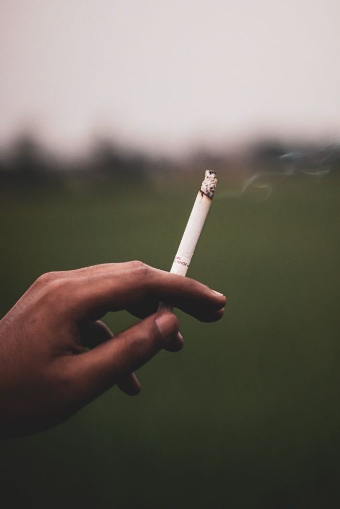 Close-up of a hand holding a lit cigarette with blurred outdoor background.